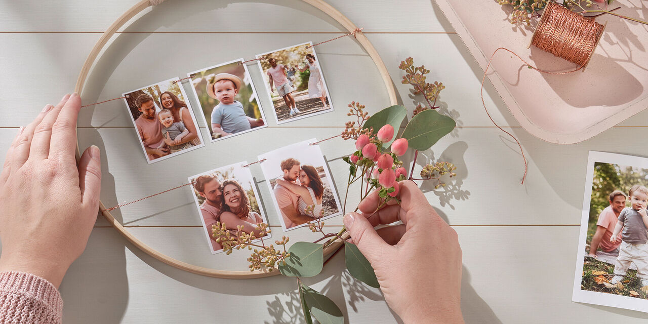 Due mani appoggiano dei fiori al lato inferiore del cerchio di legno. A fianco si vedono dello spago, fiori e una foto stampata.