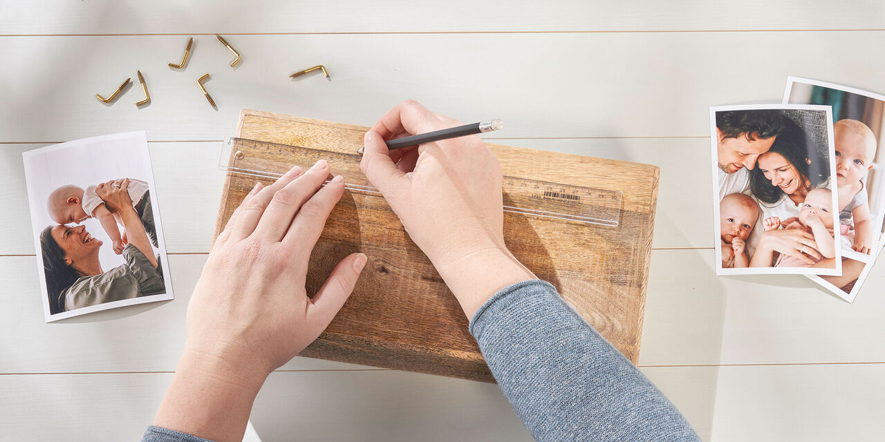 Dettagli di due mani che segnano un punto a matita su un blocco di legno utilizzando un righello. A fianco sono sparse delle stampe foto e ganci a vite.