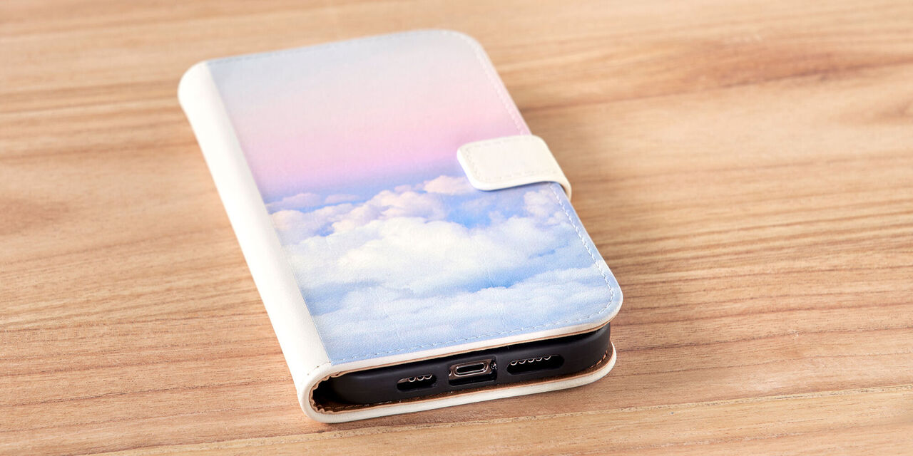 Uno smartphone è su un tavolo. Puoi vedere la custodia del telefono con un'immagine di nuvole rosa su di essa. La custodia del telefono è una tasca a ribalta laterale in pelle bianca.