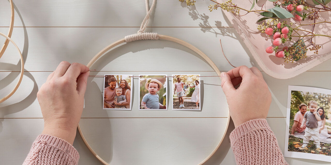 Due mani annodano un filo di spago a cui sono appese delle fotografie ai lati di un cerchio in legno. Sullo sfondo si vedono altri cerchi in legno, fiori e una foto stampata.