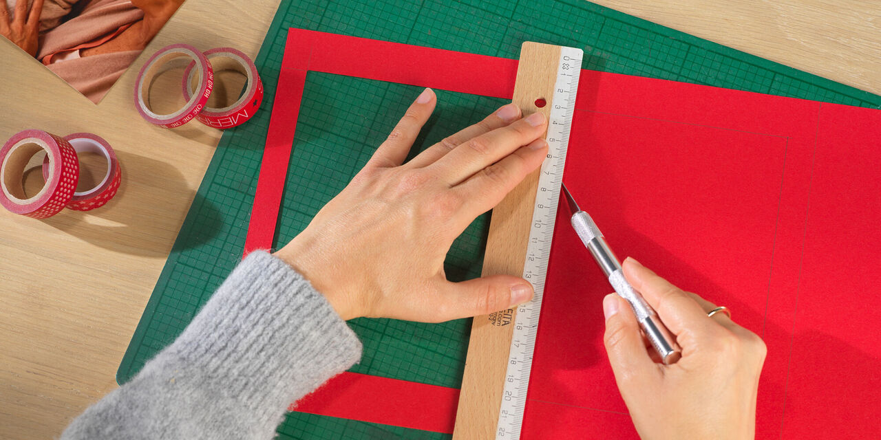 Due mani tengono un righello e una penna. Una delle quattro finestre da misurare in questo modo è già stata ritagliata nella scatola di cartone rossa.