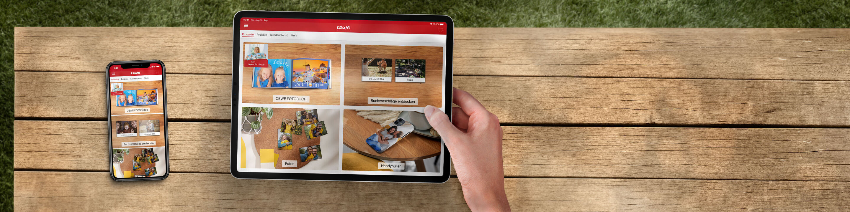 Eine Hand hält ein Tablet, auf dem CEWE FOTOBUCHs gezeigt werden, neben einem Smartphone auf einem Holztisch
