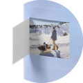 Ein Mann zieht einen Freund auf einem Schlitten durch den Schnee. Im Hintergrund sieht man verschneite Berge unter blauem Himmel. Es ist ein fröhlicher Wintertag.